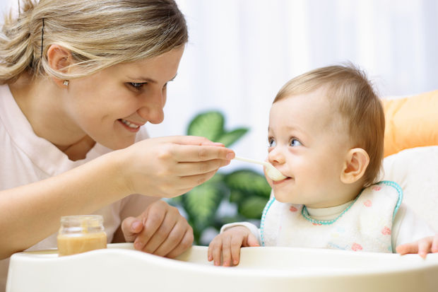 Mother feeding smiling baby with a spoon during mealtime; jar of baby food on the tray nearby.